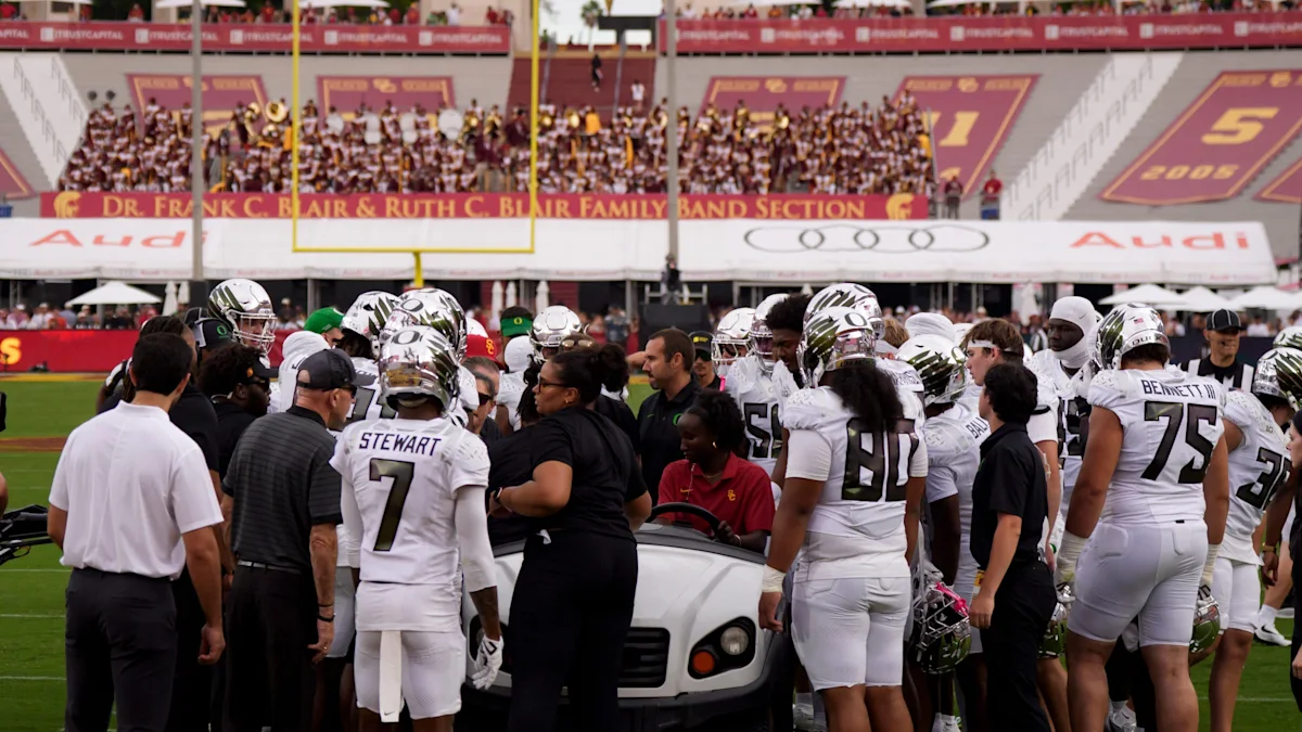 Football players and staff gather around a cart on the sideline as spectators watch from the stands.