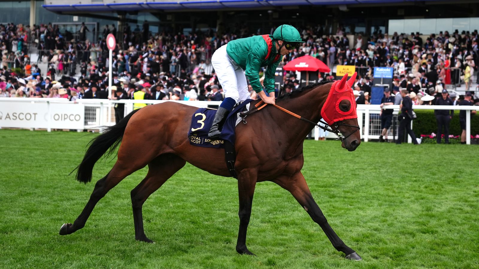 Racehorse wearing red blinkers carries jockey in green silks across a grass paddock.