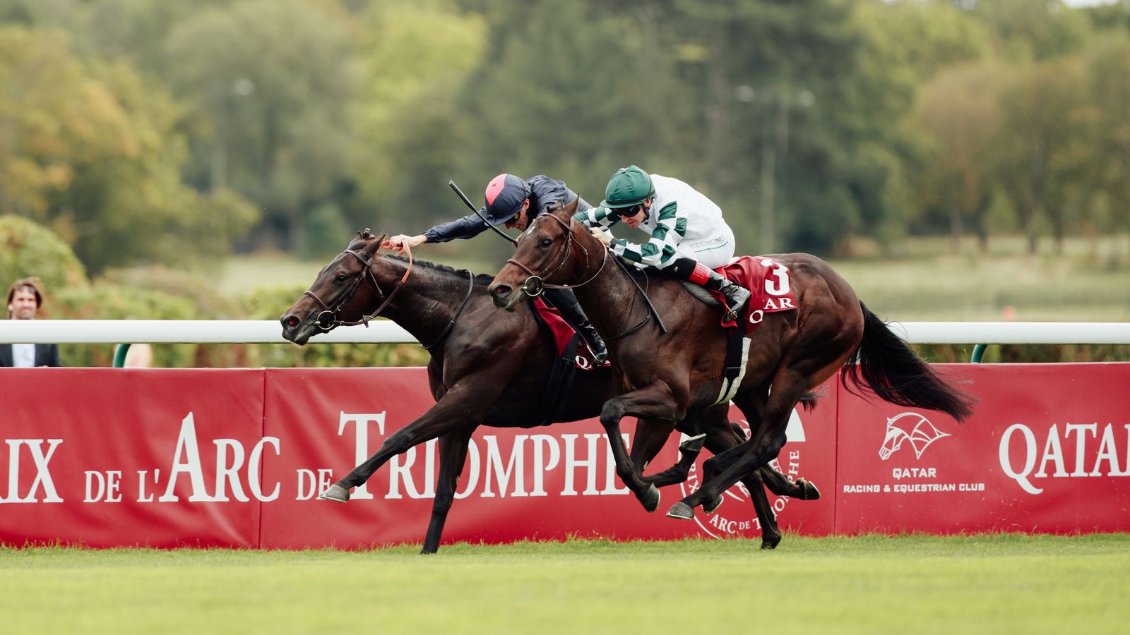 Two jockeys race dark brown horses past red Prix de l’Arc de Triomphe barriers