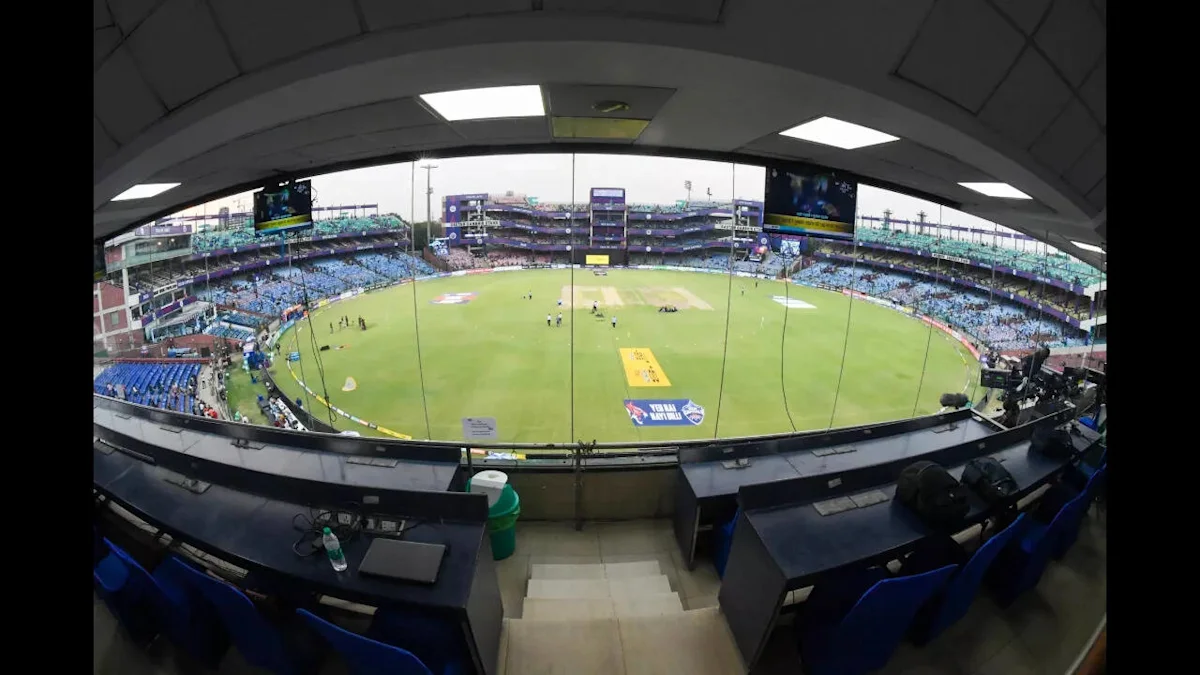 Wide view of a cricket stadium through glass, with the field, stands, broadcast desks and empty blue chairs.