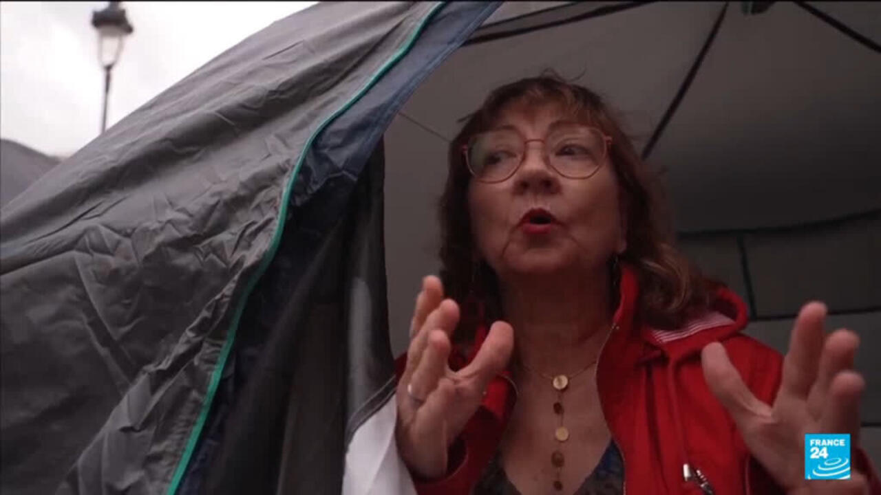 Woman in glasses and a red jacket gestures from inside a tent.