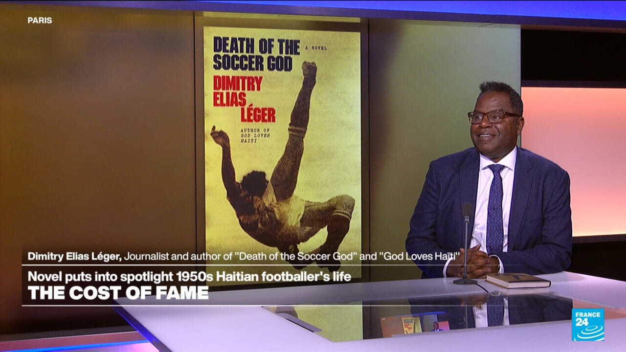 A suited man with glasses sits beside a displayed book cover showing a soccer player kicking a ball.