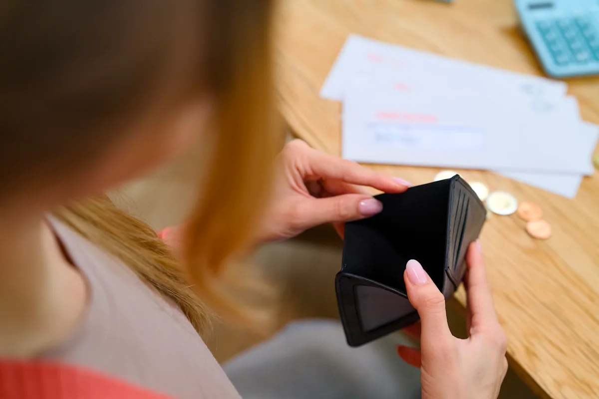 Person examines an empty black wallet beside bills and coins on a wooden table