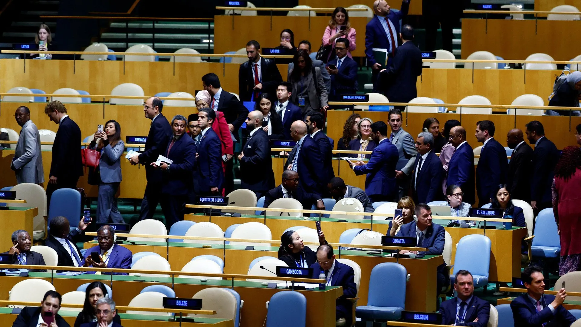 Delegates in formal attire move through rows of seats in a large assembly chamber.