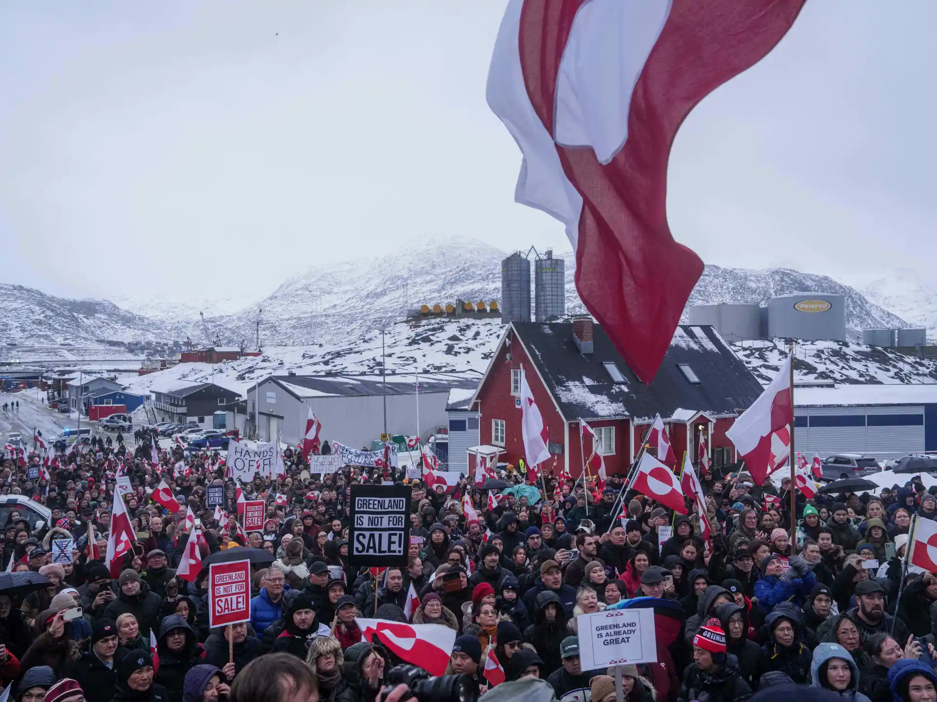 Crowd gathers beneath Danish and Greenlandic flags in a snowy town with mountains behind