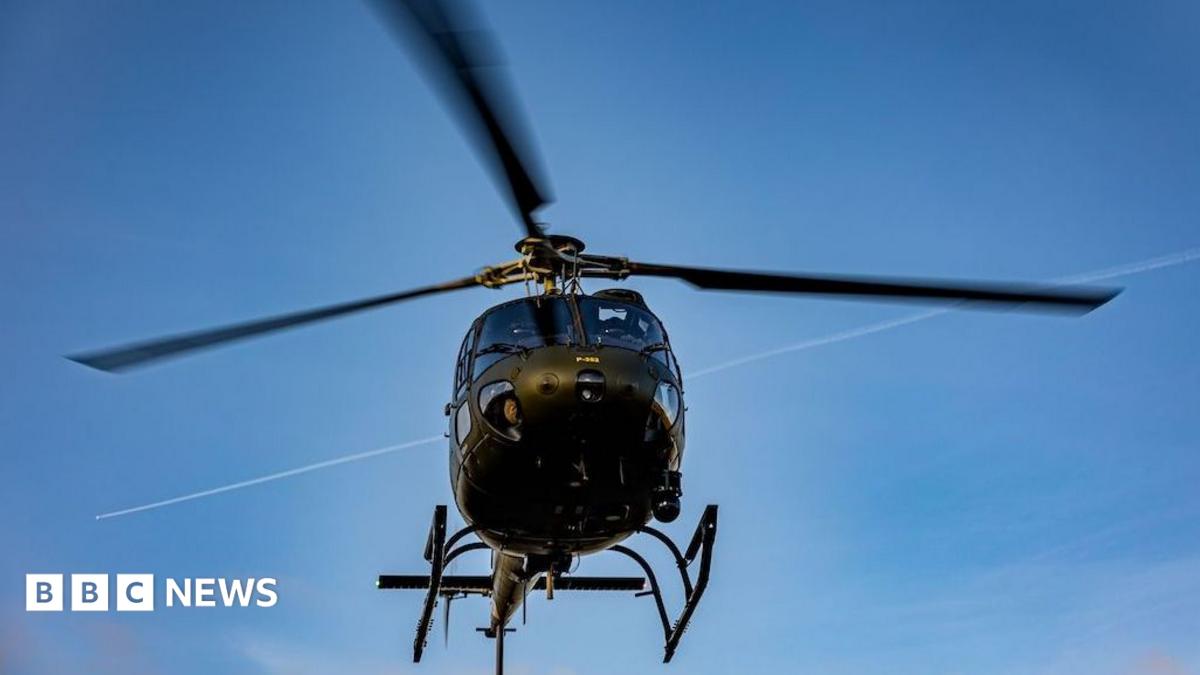 Military helicopter viewed from below against a blue sky, with blurred rotor blades overhead.
