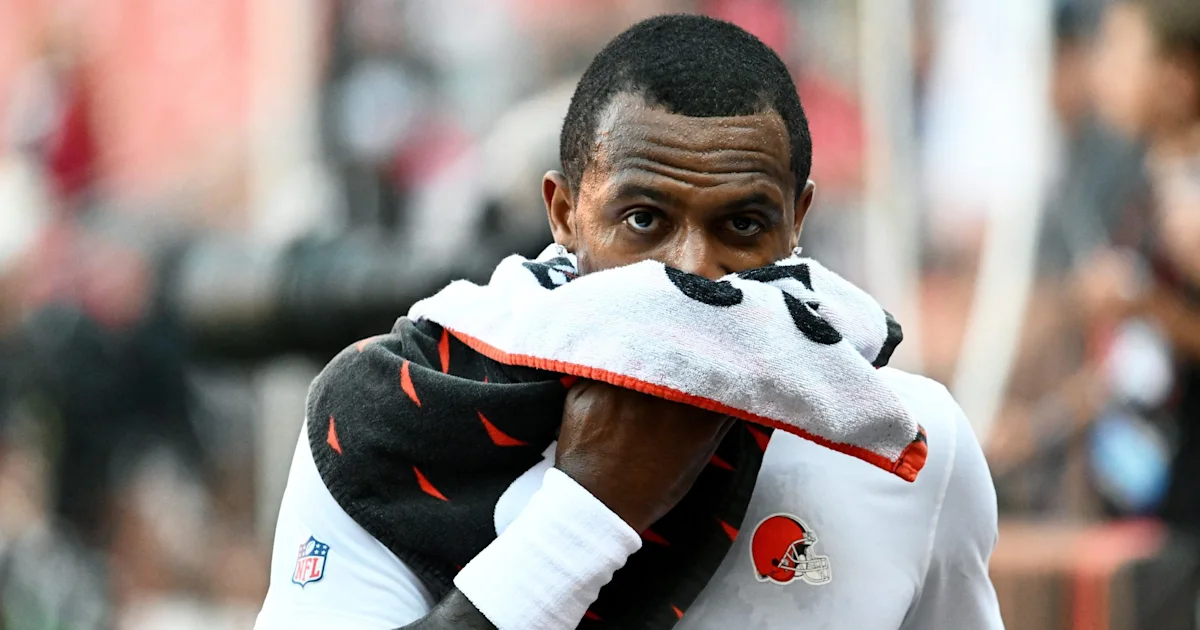 Football player holds a towel over his mouth, with a blurred crowd behind him.