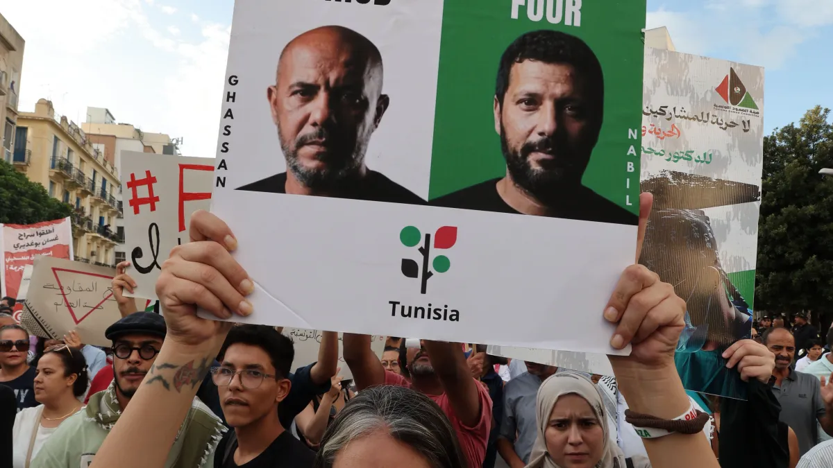 Protesters hold a Tunisia solidarity poster featuring portraits of two men above a crowd in the street.