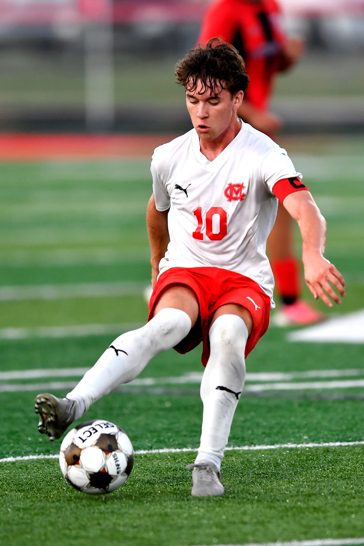 Soccer player in white jersey and red shorts strikes a ball on an artificial turf field.