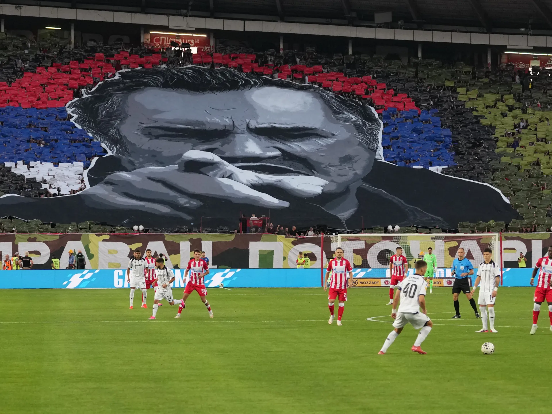 Football match underway before a stadium display featuring a large monochrome portrait and colored crowd sections.