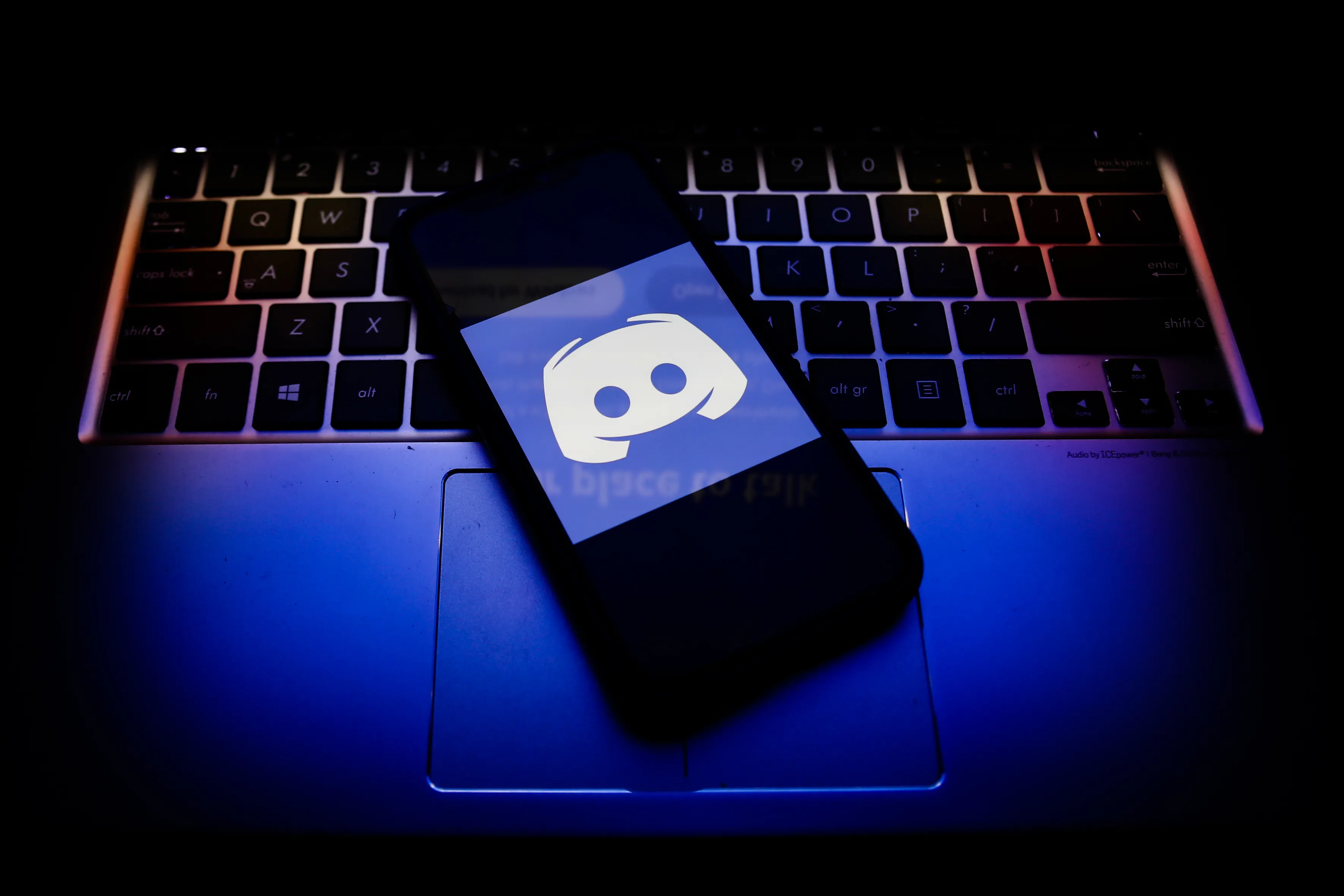 A smartphone displaying the Discord logo rests on a blue-lit laptop keyboard.