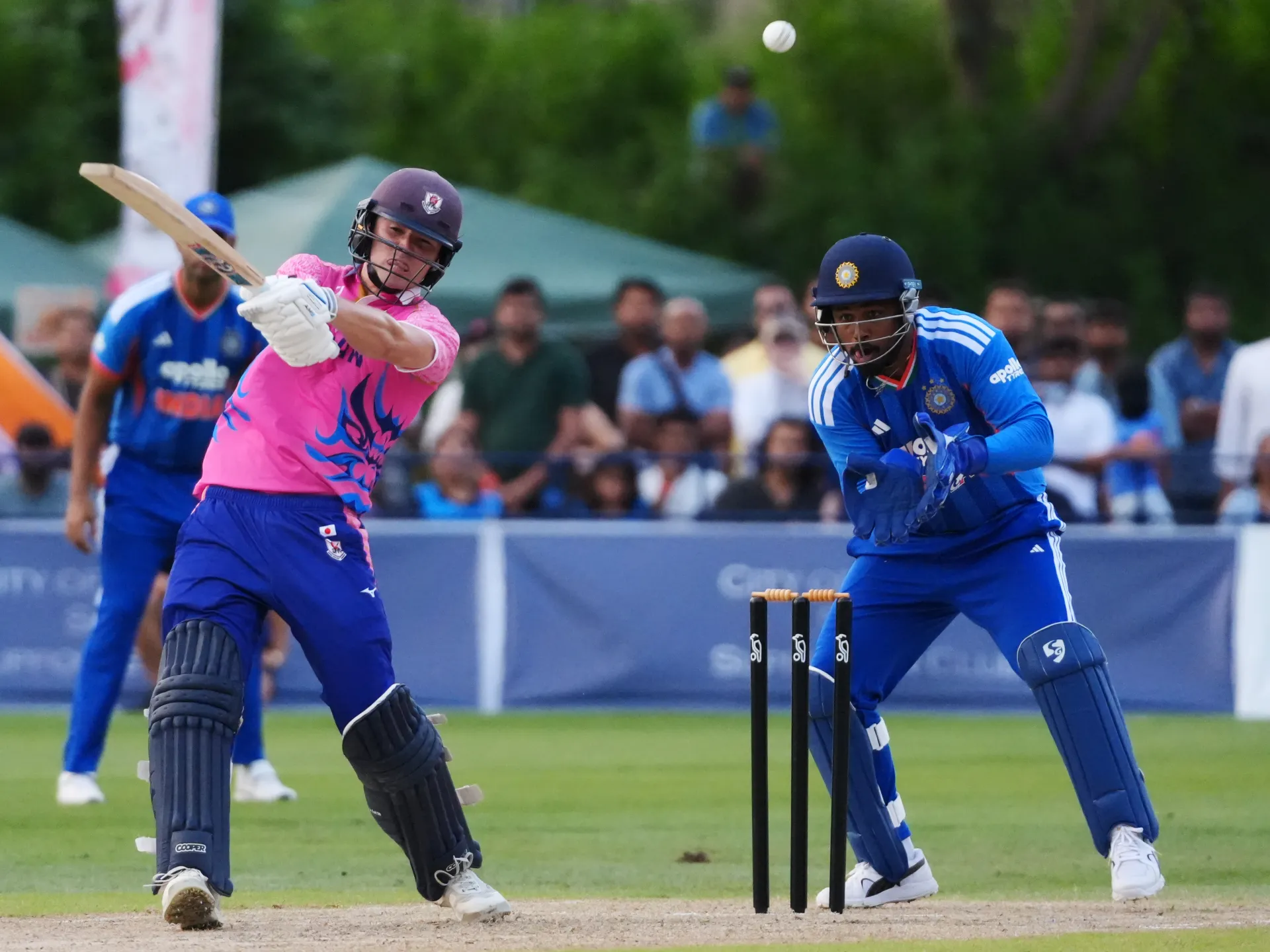 Cricketer in pink swings at a ball as a blue-clad wicketkeeper crouches behind stumps.