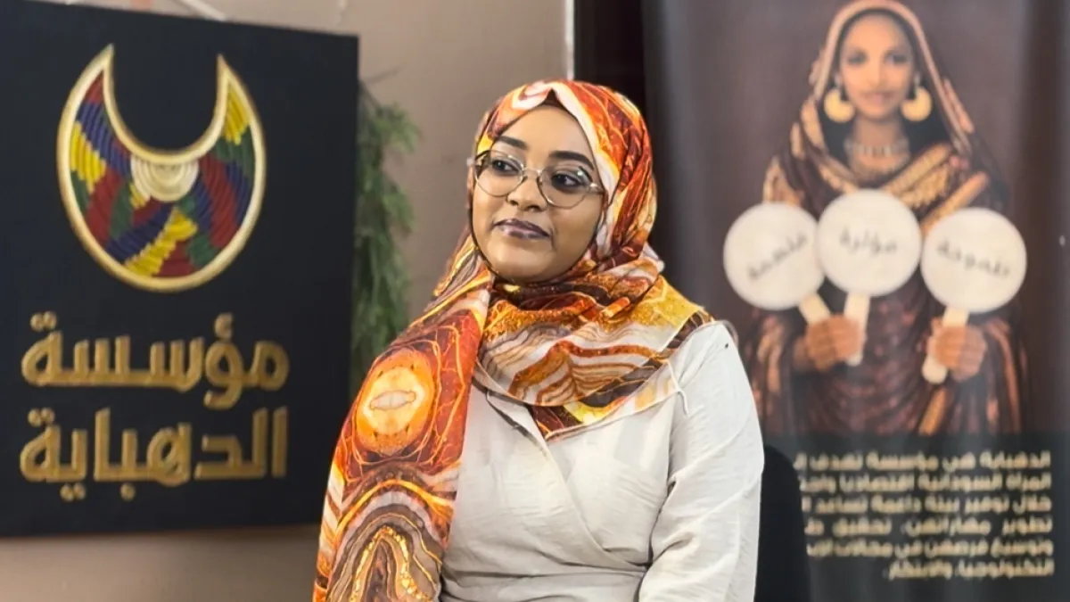A woman in glasses and a colorful headscarf stands between two Arabic-language display banners.