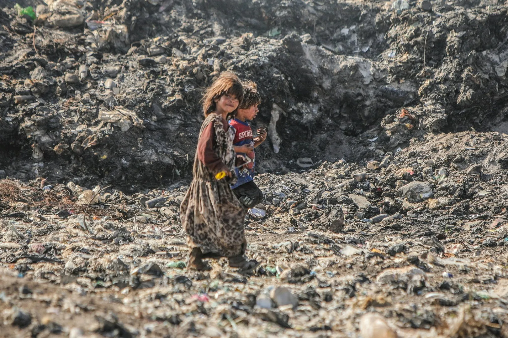 Two children walk through a large hillside of scattered garbage, with one carrying a sack.