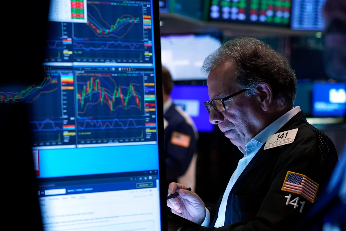 Trader studies market charts on multiple monitors while holding a pen on a busy trading floor.