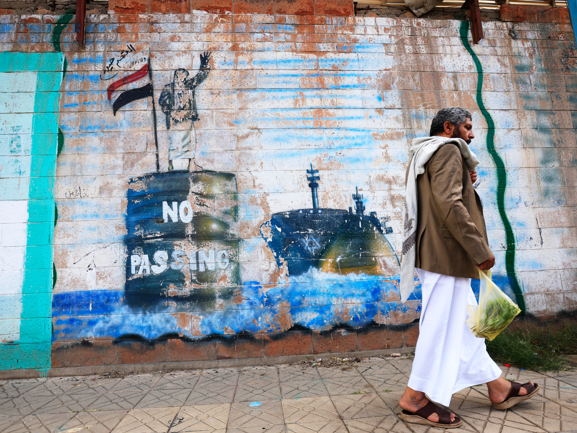Man in traditional clothing walks past a weathered, graffiti-covered brick wall carrying a bag of vegetables.