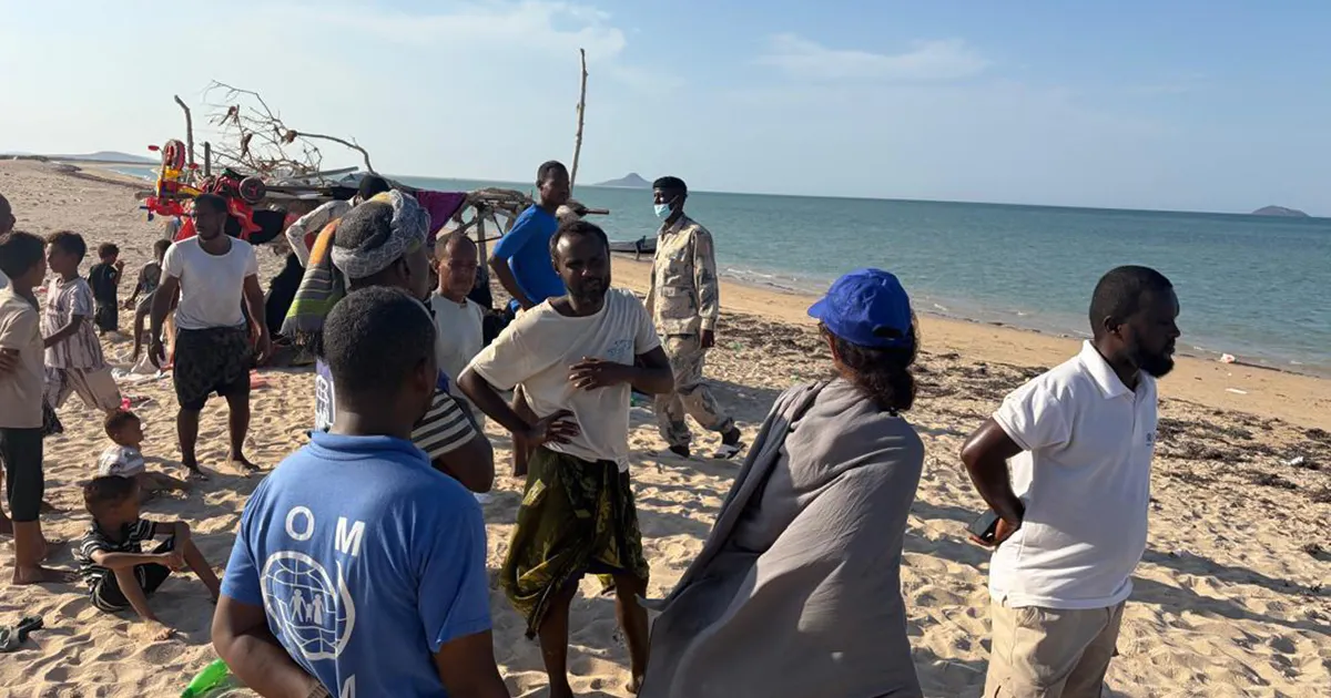 People gather on a sandy beach beside the sea, with boats and an aid worker visible.