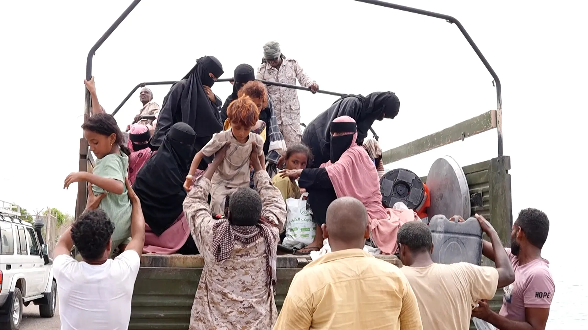 Children and adults crowd onto an open military truck beside uniformed personnel.