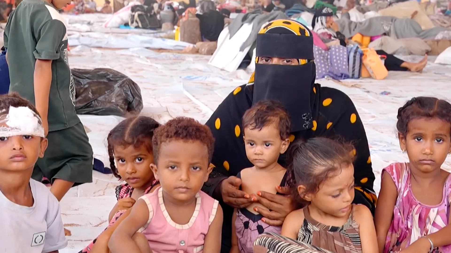 Children sit with a veiled woman in a crowded shelter filled with blankets and belongings.