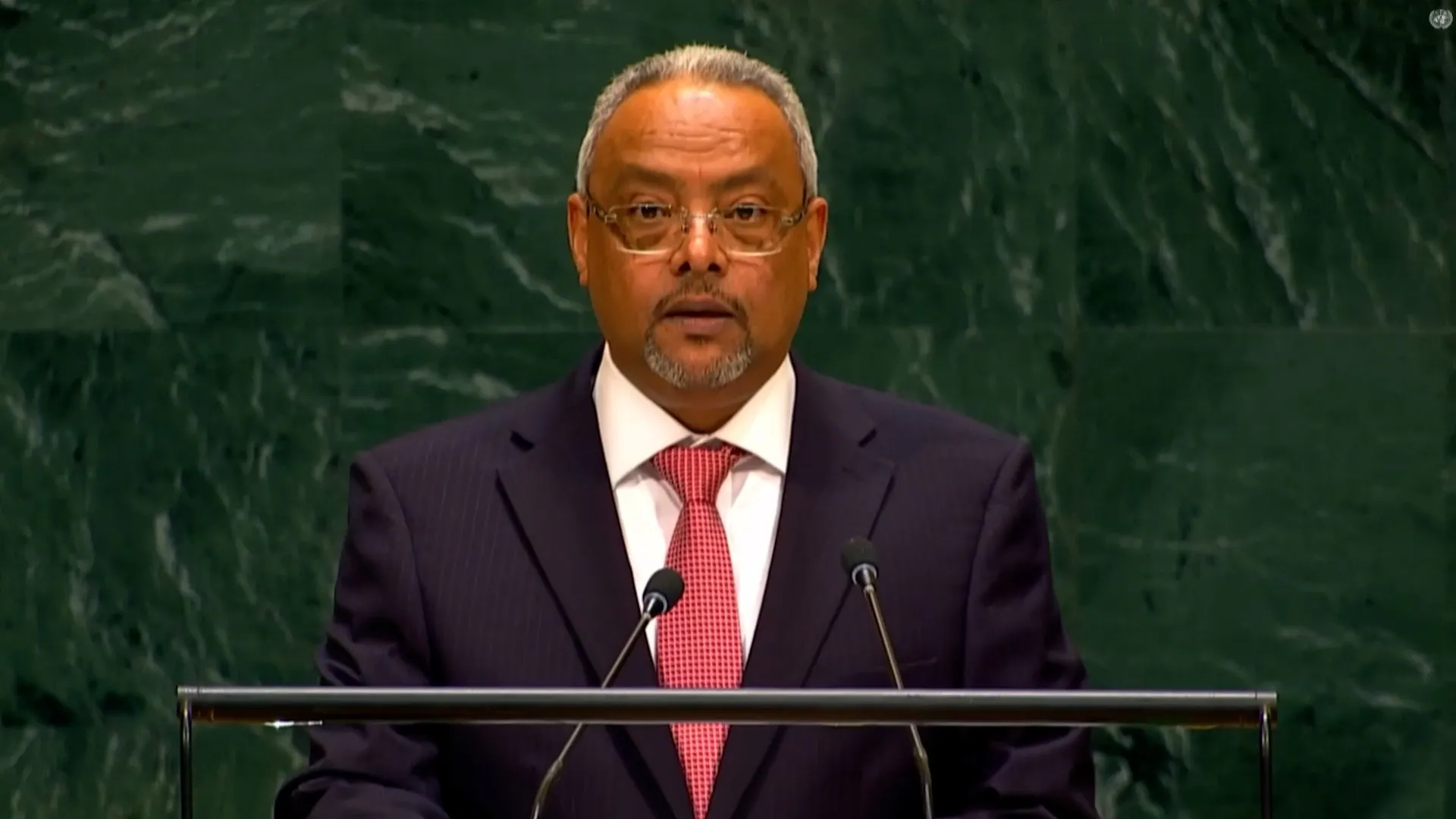A gray-haired man in glasses speaks at a lectern with two microphones against a green marble wall.