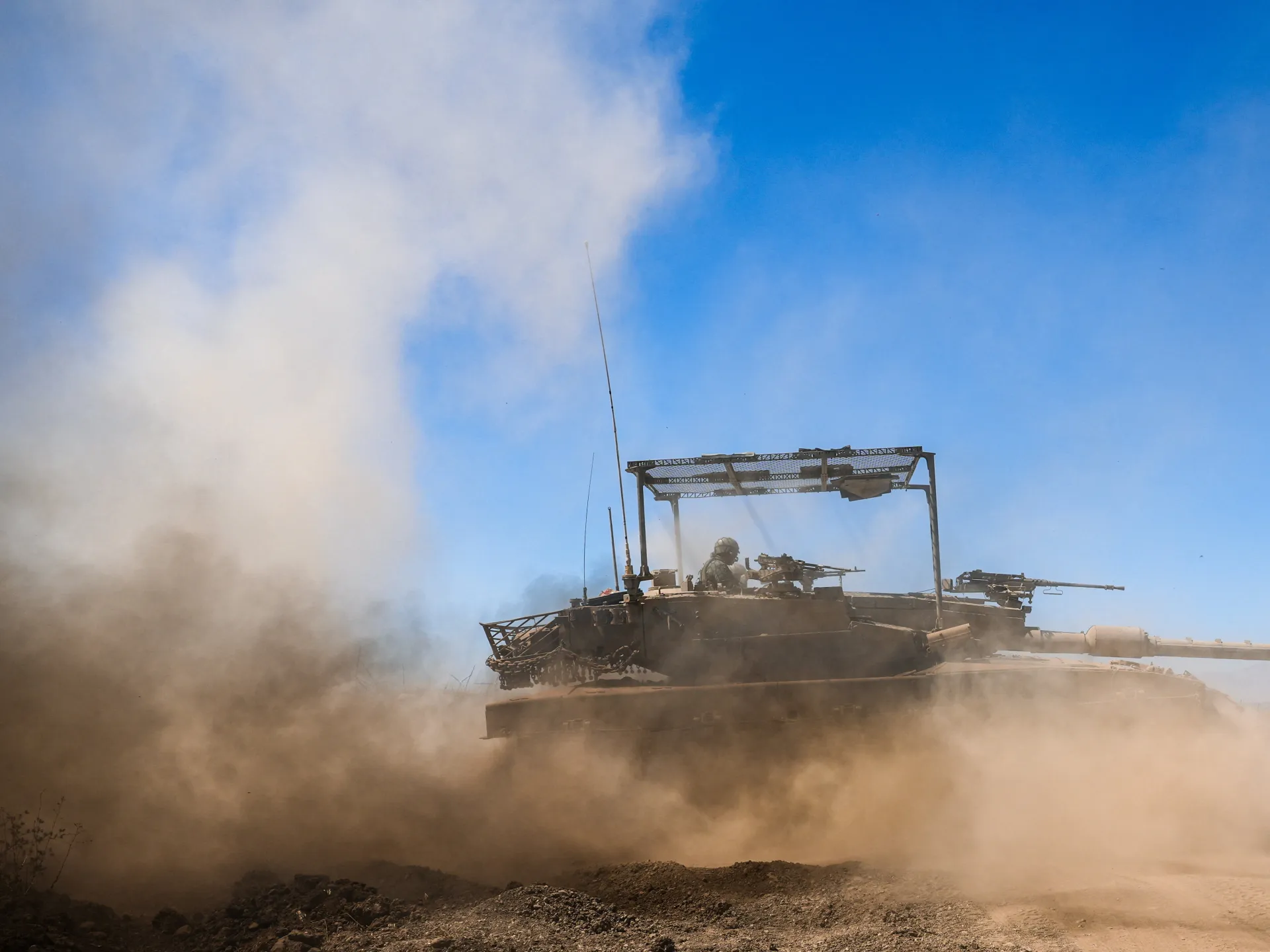 Dust-covered tank advancing across rough terrain beneath a bright blue, partly clouded sky.