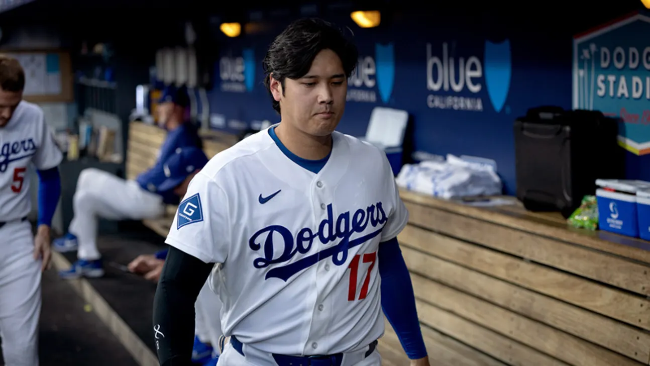 Baseball player in Dodgers uniform walking through a dugout while teammates sit in the background