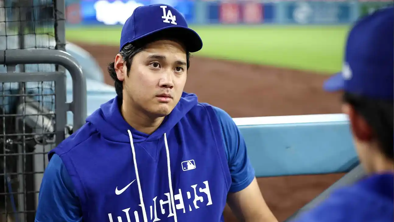 Baseball player in Dodgers cap and blue hoodie sits near a dugout during a game.