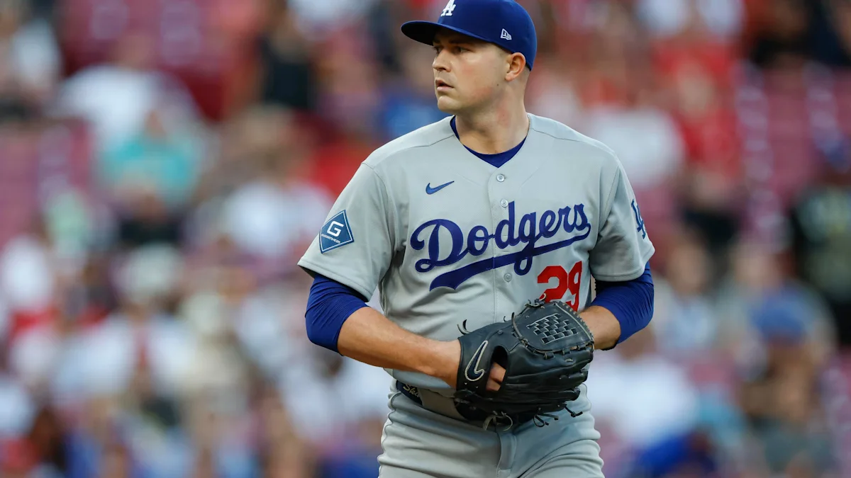 A Dodgers pitcher in a gray uniform and blue cap holds a glove on the field.