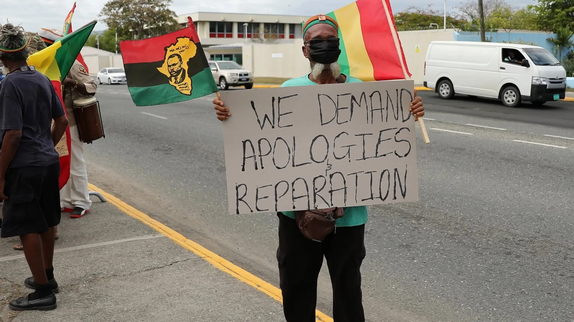 Masked protester holds a sign demanding apologies and reparations beside flags and fellow demonstrators.