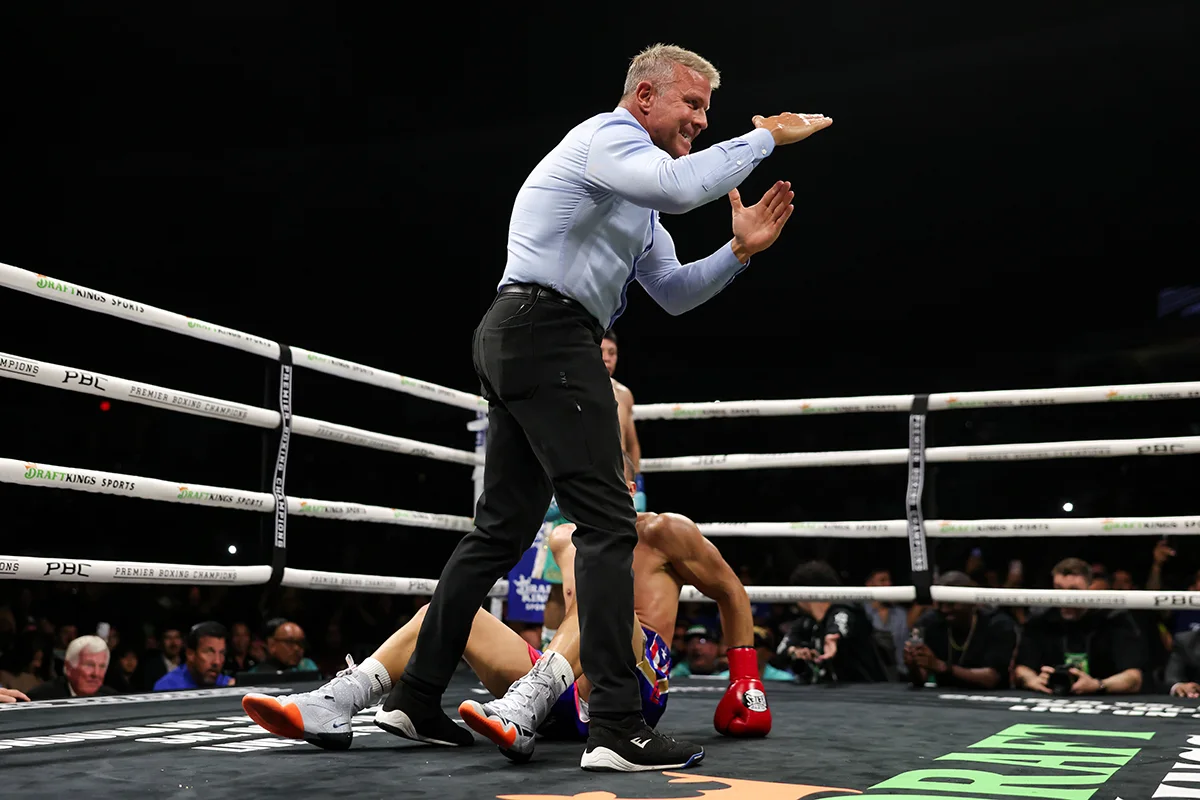 Referee gestures over a seated boxer in the ring as another fighter lies nearby after a knockdown.