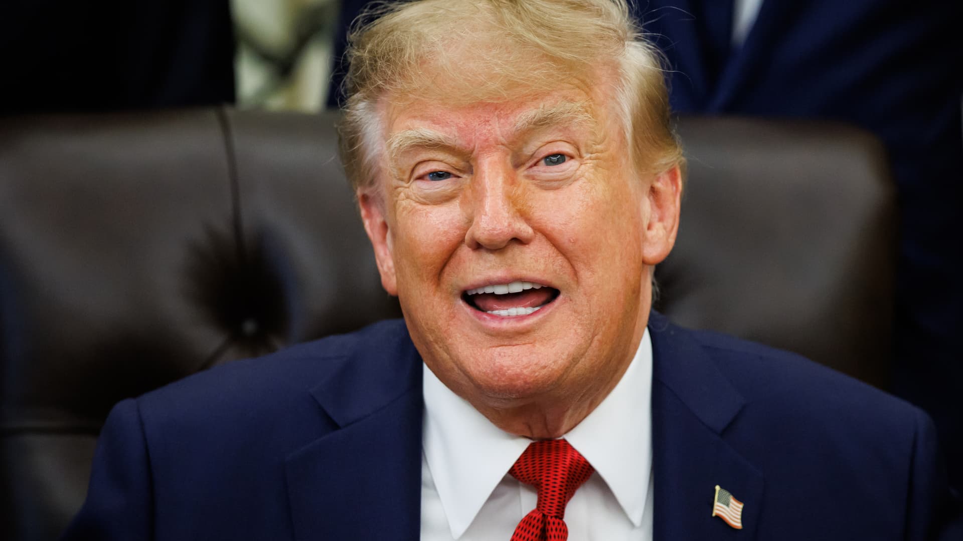 Donald Trump speaks toward the camera, wearing a navy suit, white shirt, and red tie.