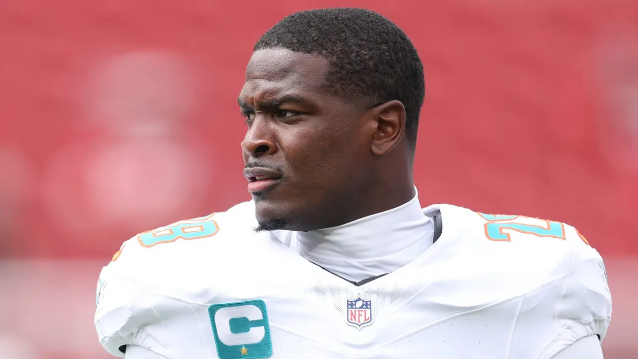 A Miami Dolphins player in a white uniform looks to the side against a blurred red background.