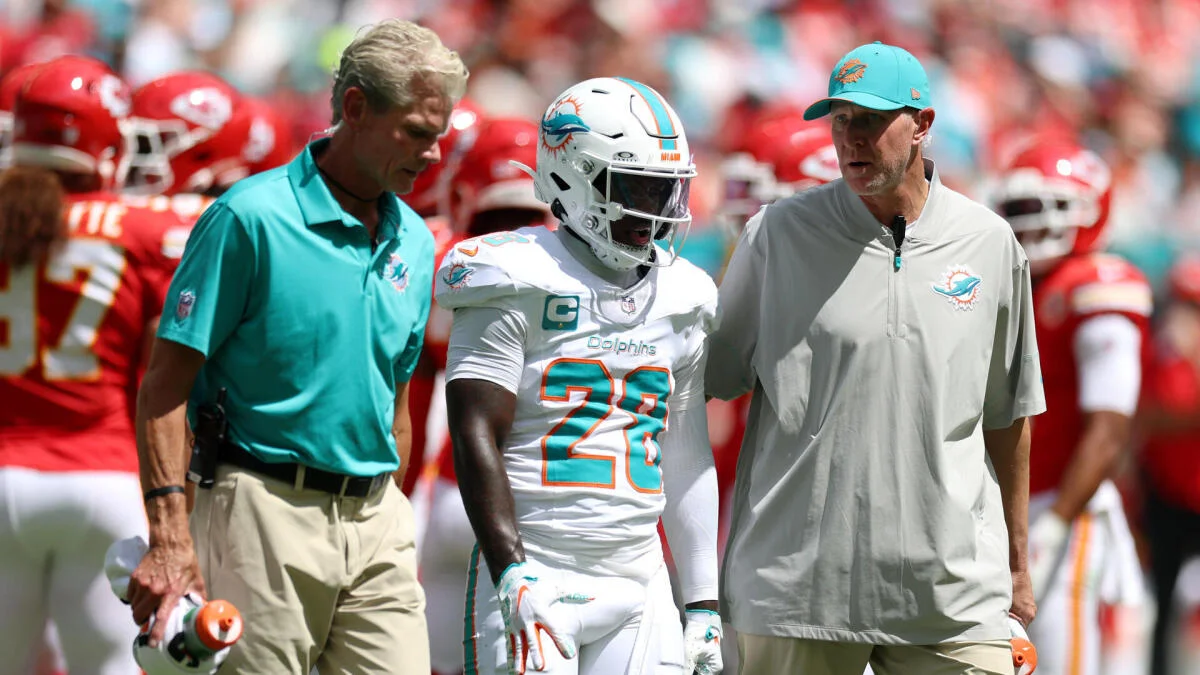 A Dolphins player in a white uniform walks between two team staff members on the sideline.