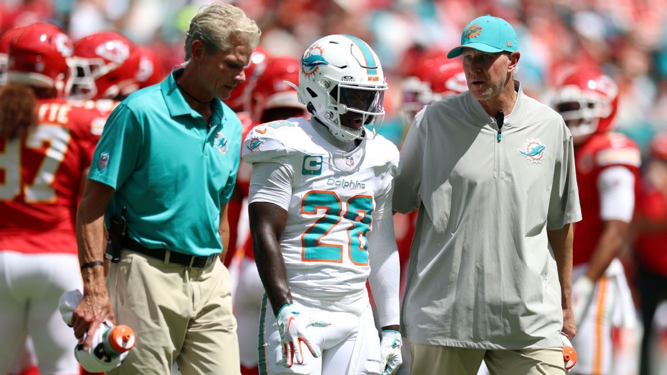 Dolphins player in a white uniform walks between two staff members on a football field.