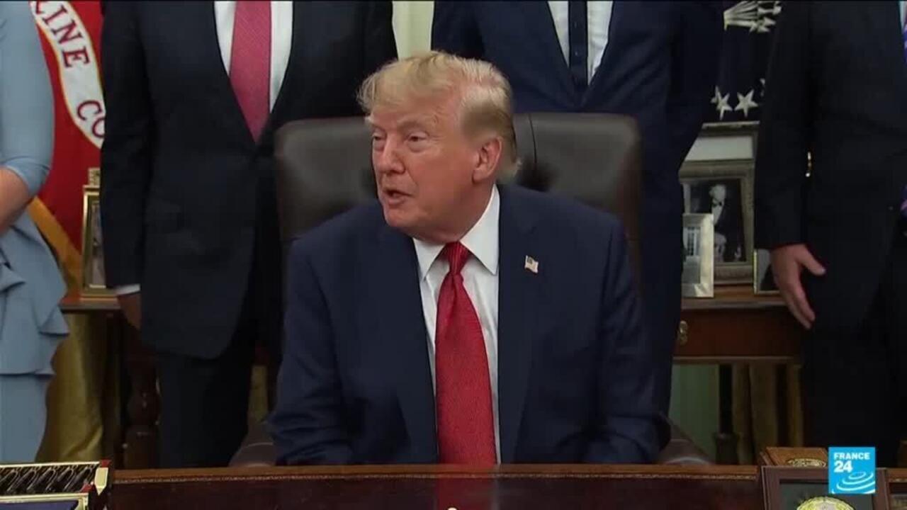 Donald Trump sits at a desk in a suit, with officials standing behind him.