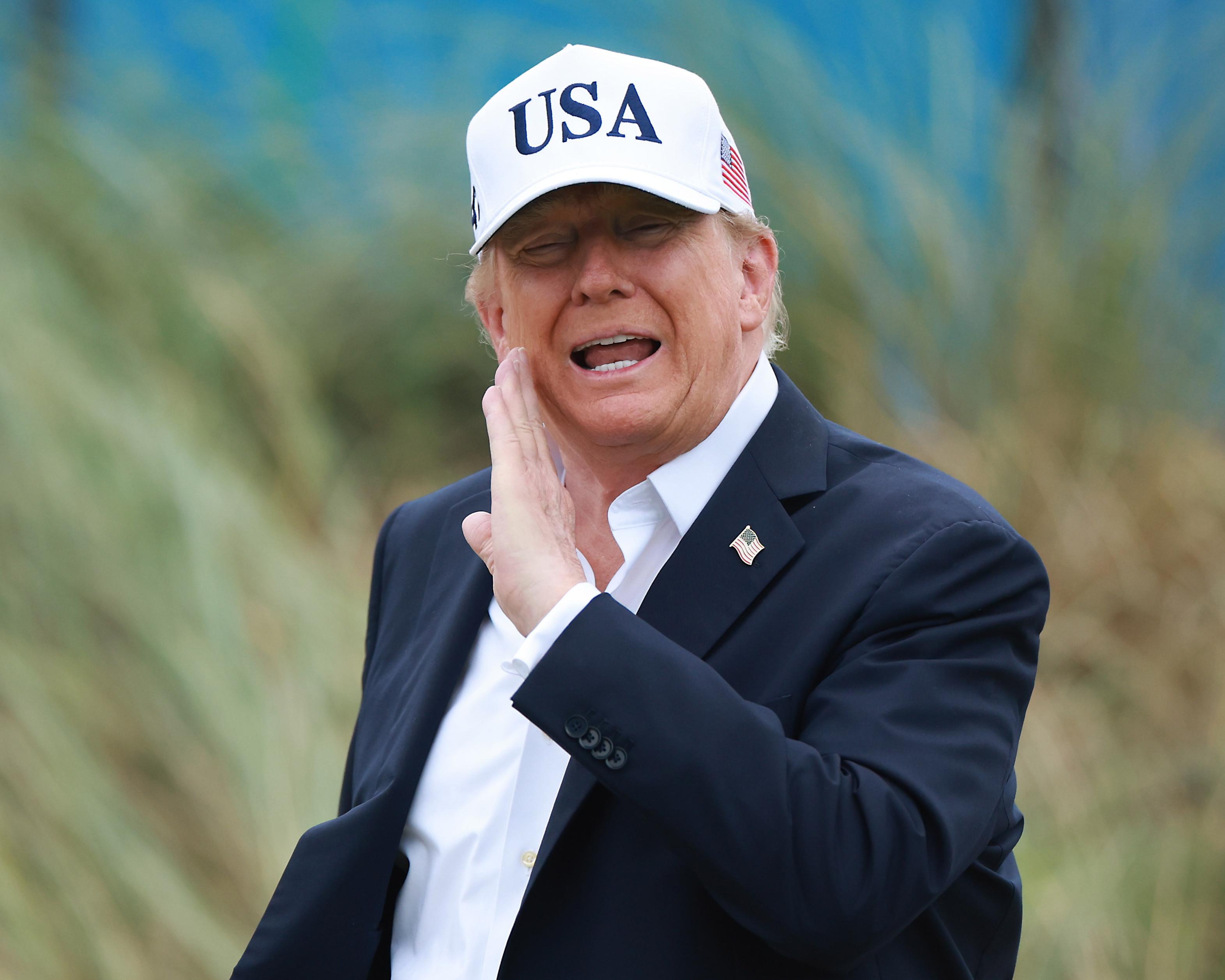 Donald Trump wearing a USA cap and suit, raising one hand near his face.