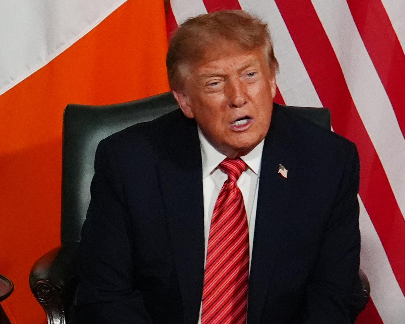 Donald Trump seated in a dark suit and red tie before American and Irish flags