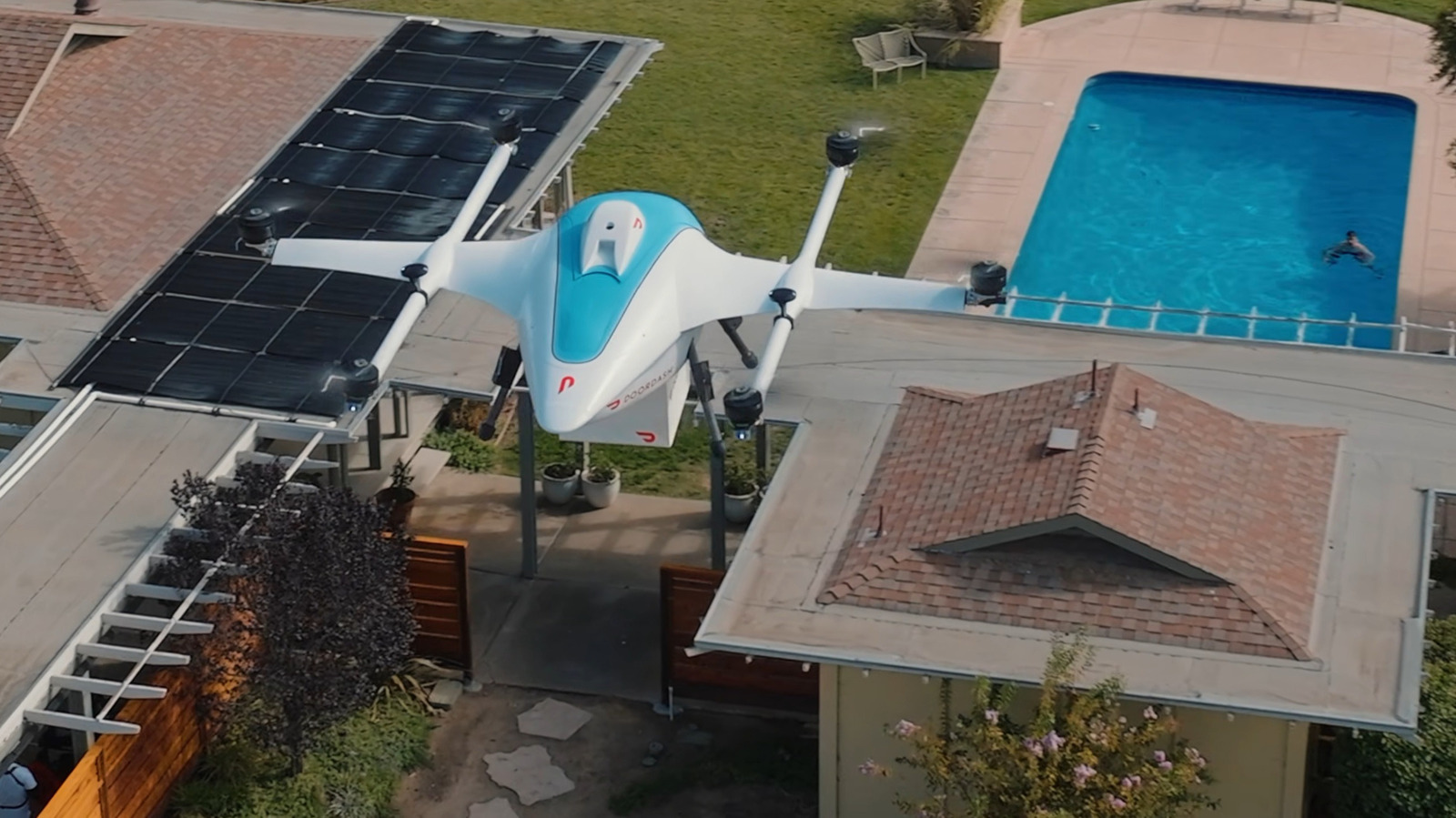 White delivery drone with a blue canopy hovers above a residential patio beside solar panels and a swimming pool.
