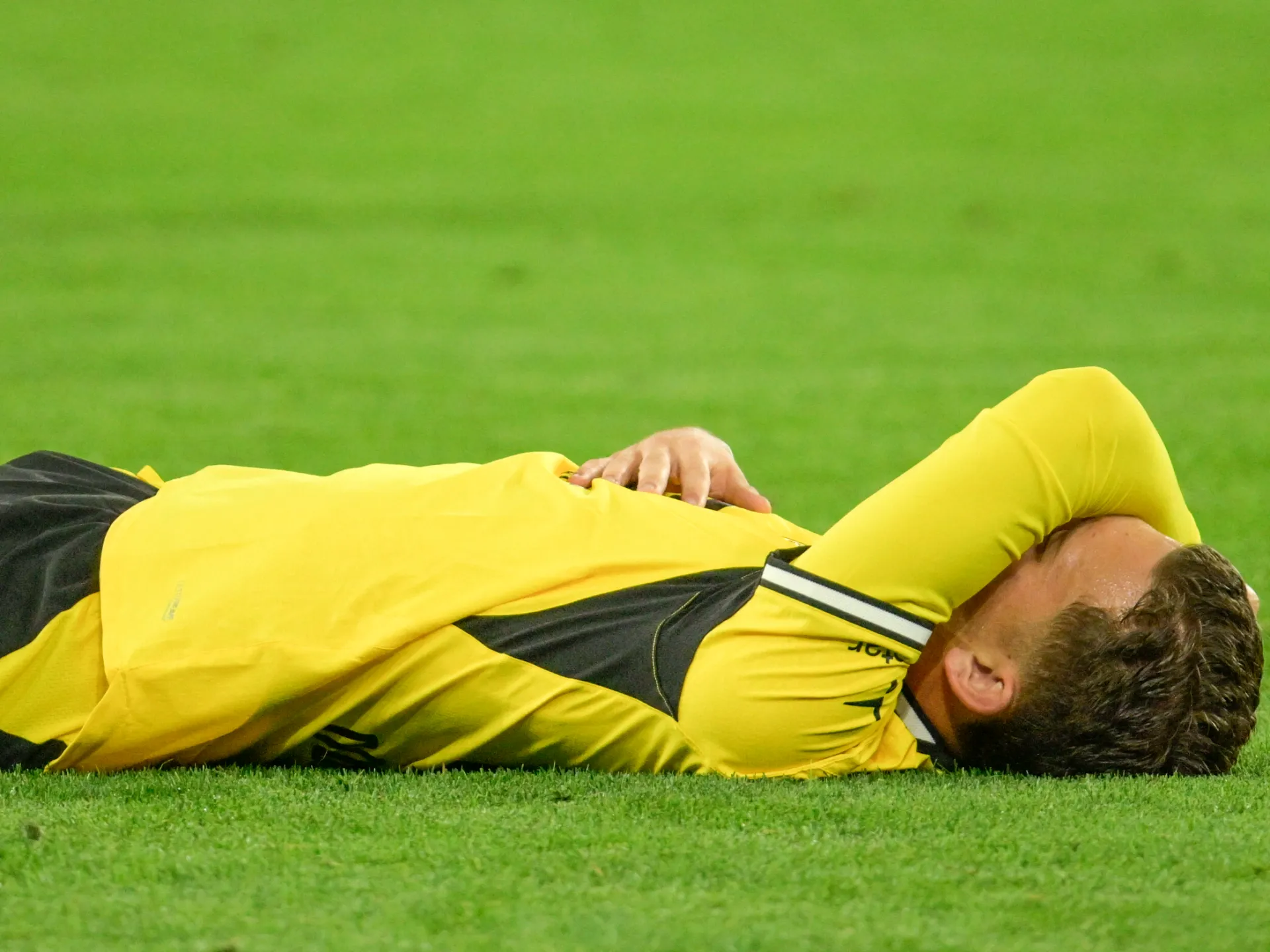 Football player in yellow kit lies on his side on the grass, appearing injured during a match.