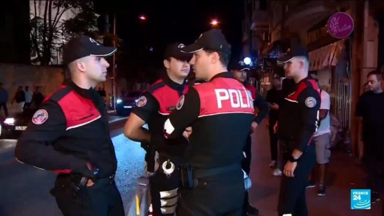 Police officers in red and black uniforms stand outside a nighttime venue with bystanders nearby.