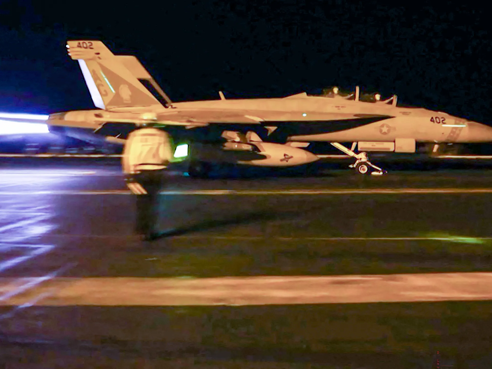 Nighttime fighter jet on a wet runway, viewed from the side with bright exhaust and external stores
