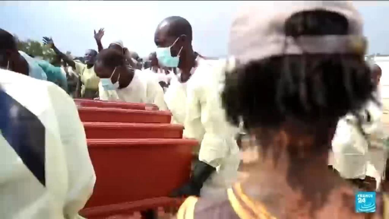 People in protective clothing carry red coffins through a crowd, with one person wearing a face mask.