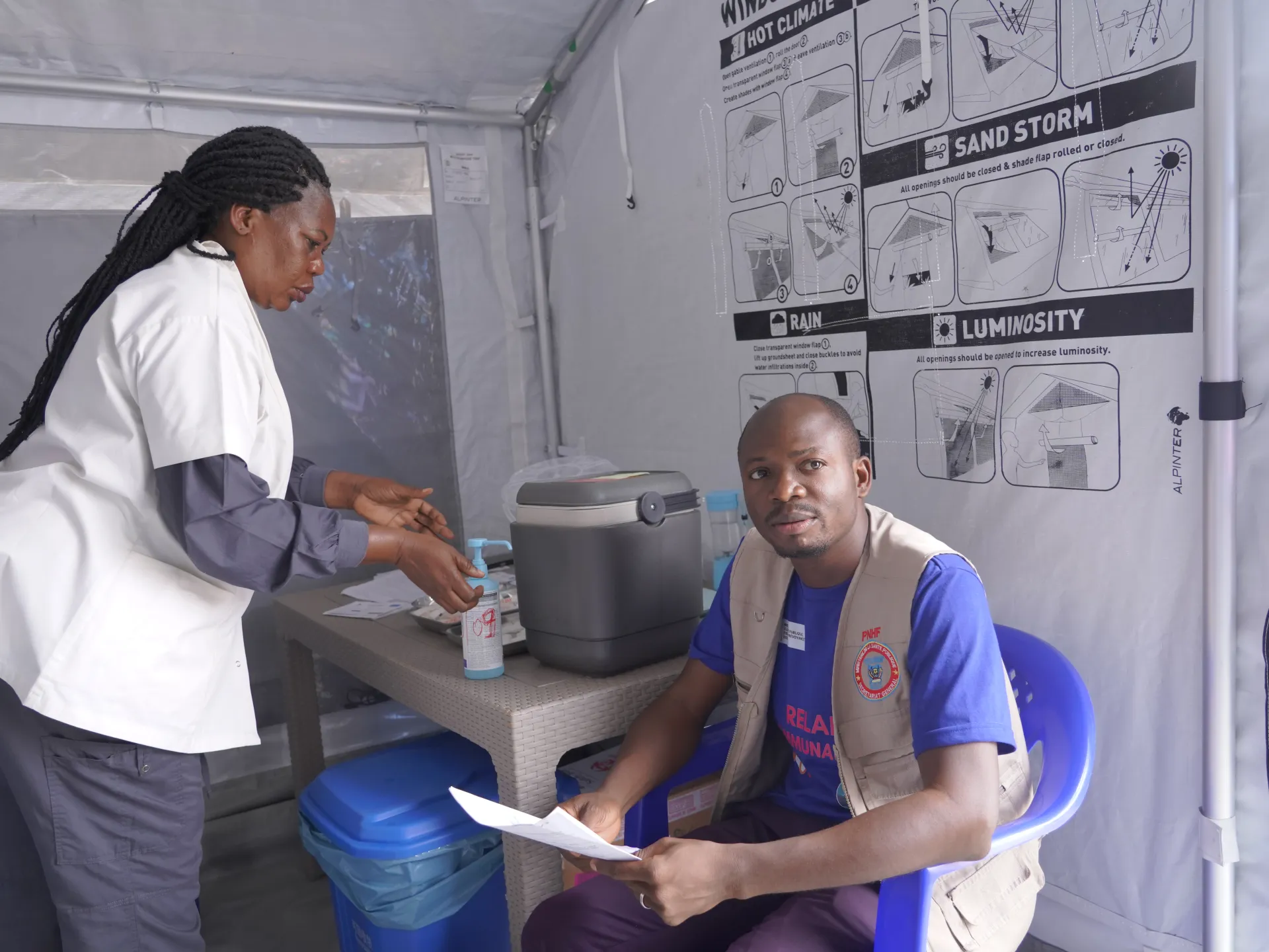 Two health workers handle medical supplies inside a temporary clinic tent.