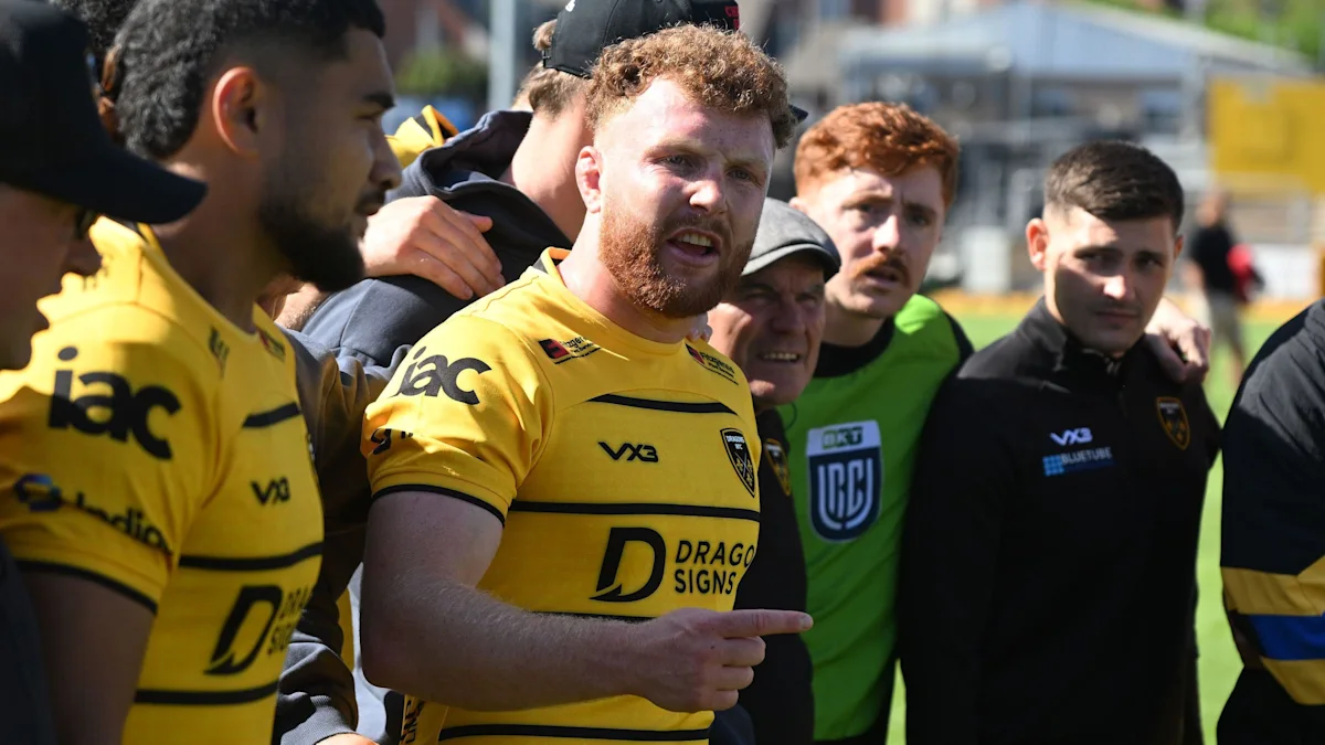 Rugby player in yellow Dragons jersey surrounded by teammates and staff during an outdoor matchday moment