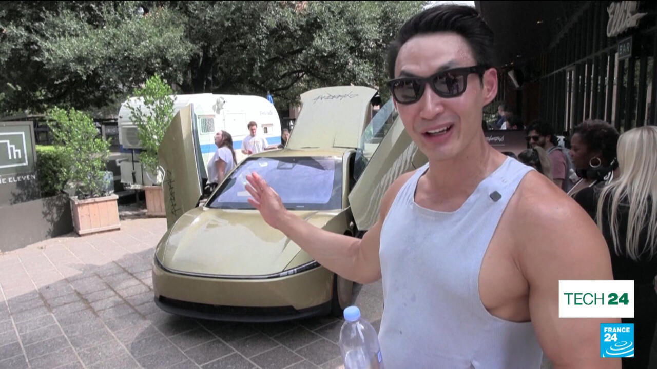 Man gestures beside a futuristic car with its doors raised outside a building.