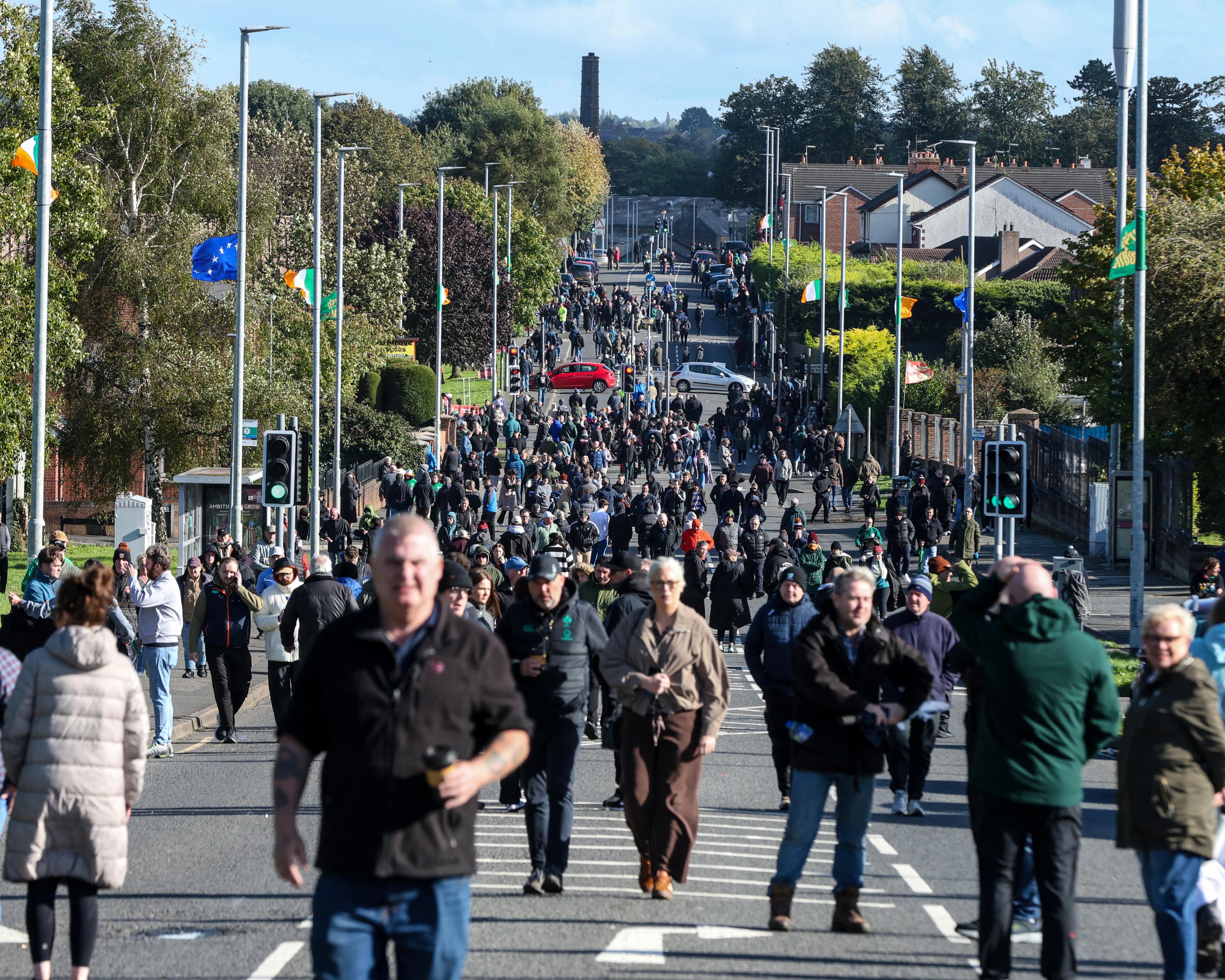 A large crowd walks along a road past traffic lights, houses, trees, and Irish flags.