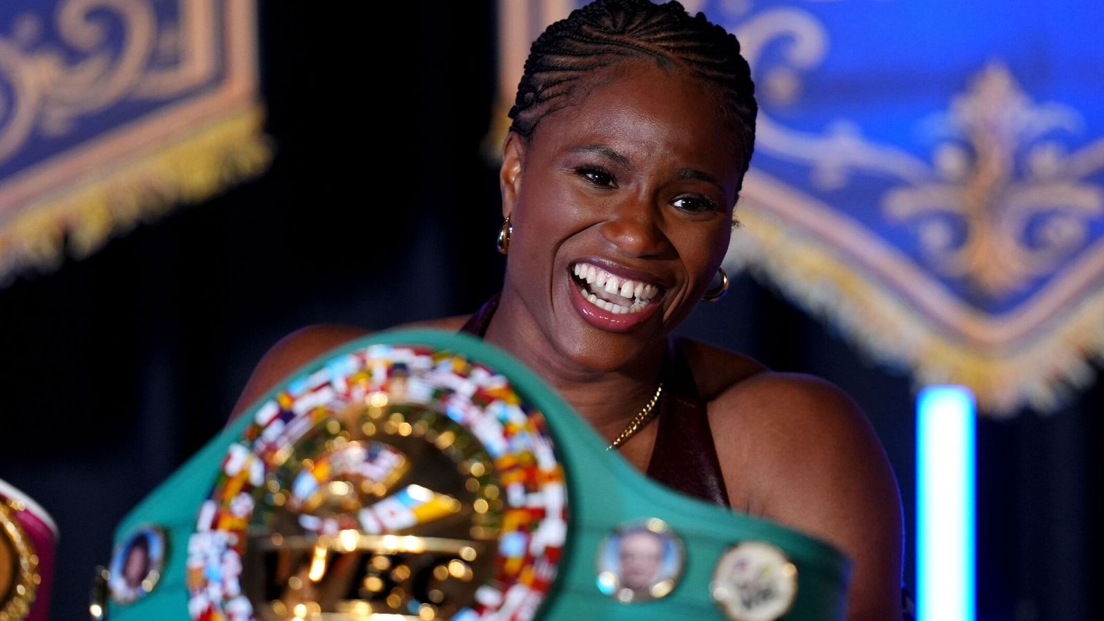 Smiling boxer with braided hair holds a colorful championship belt toward the camera.