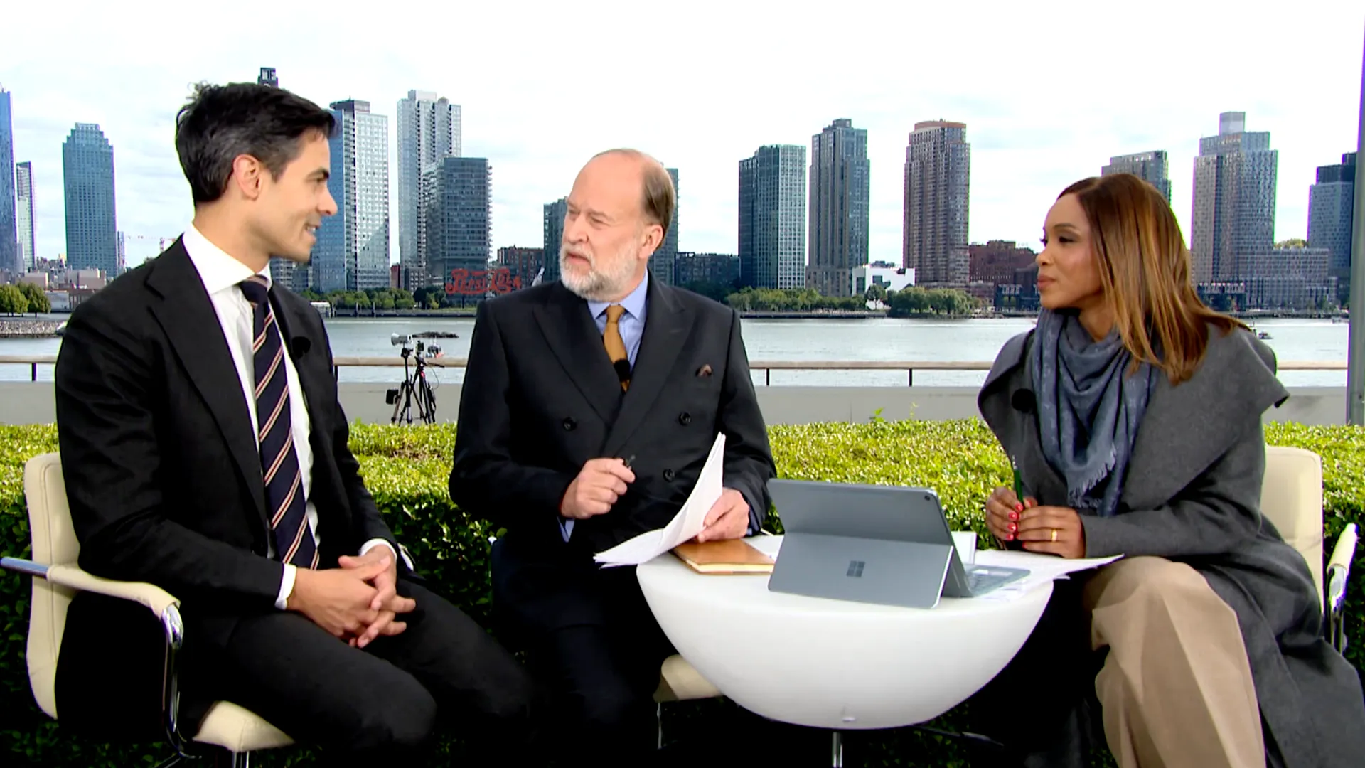 Three formally dressed people sit around a table with papers and a tablet, against a city waterfront backdrop.