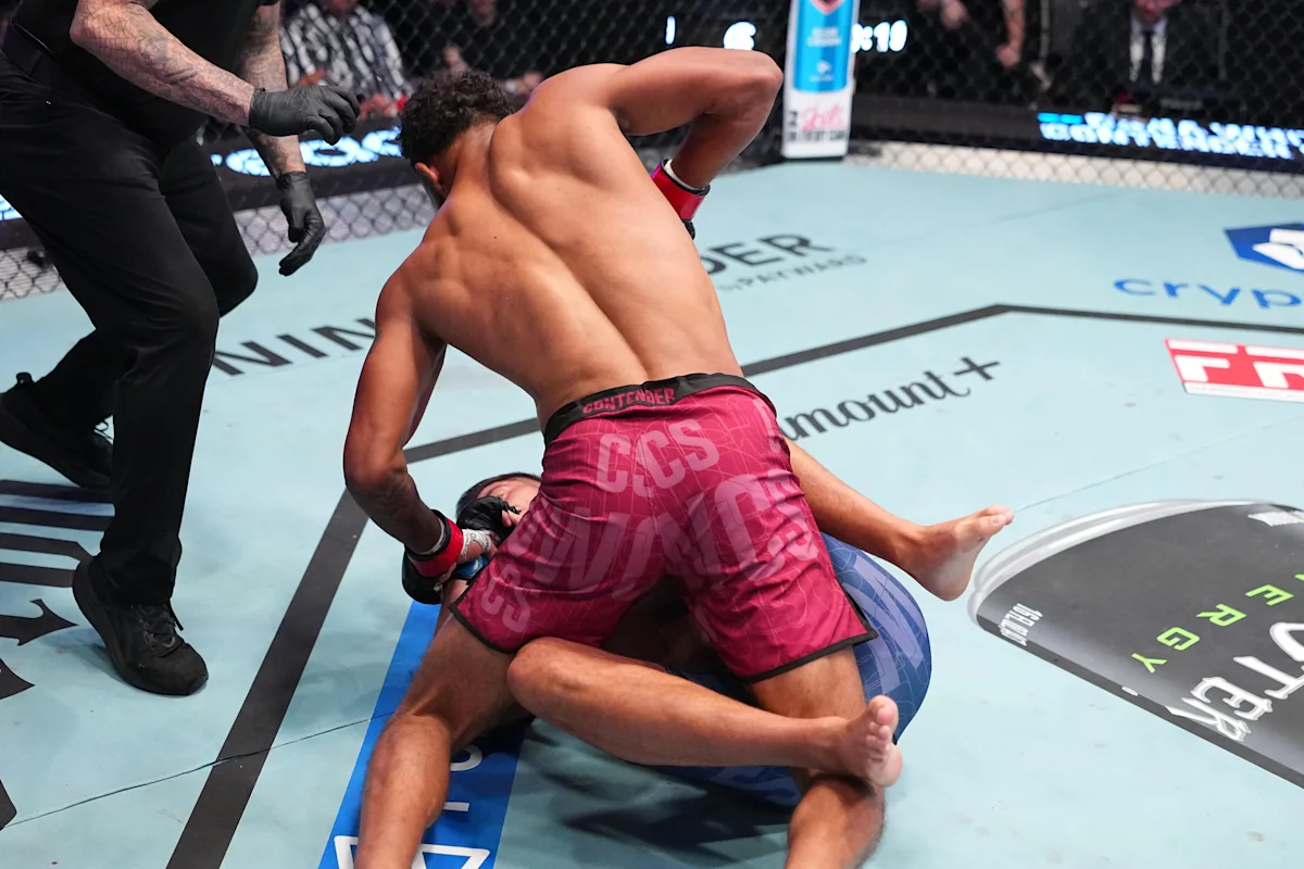An MMA fighter in pink shorts grapples with an opponent on the canvas as a referee approaches.