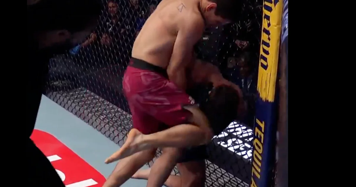 A fighter in red shorts presses an opponent against the chain-link cage during a bout.